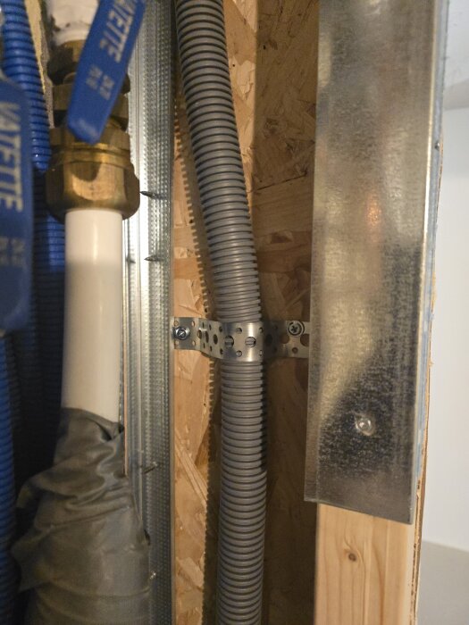 Flexrör och vattenrör monterade vid en väggkonstruktion med OSB-skiva, fästa med hålband och omgivna av trä- och metallreglar.