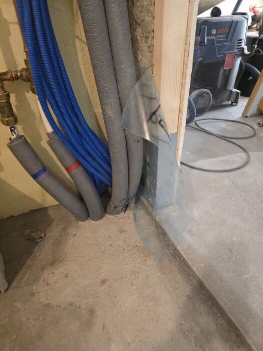 Gråa och blå flexrör vid en vägg, synligt intill en träregel. Verktyg och utrustning i bakgrunden.