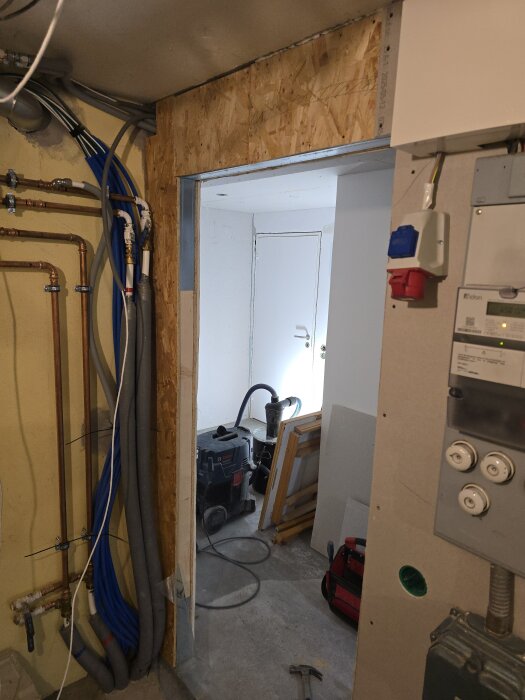 Rum under renovering med öppning i väggen, synliga rör och kablar. Byggmaterial, verktyg och en verktygslåda är placerade vid öppningen.