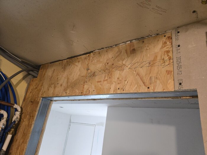 OSB-skiva över en dörröppning med synliga rör och reglar i ett hobbyrum/gym under renovering.