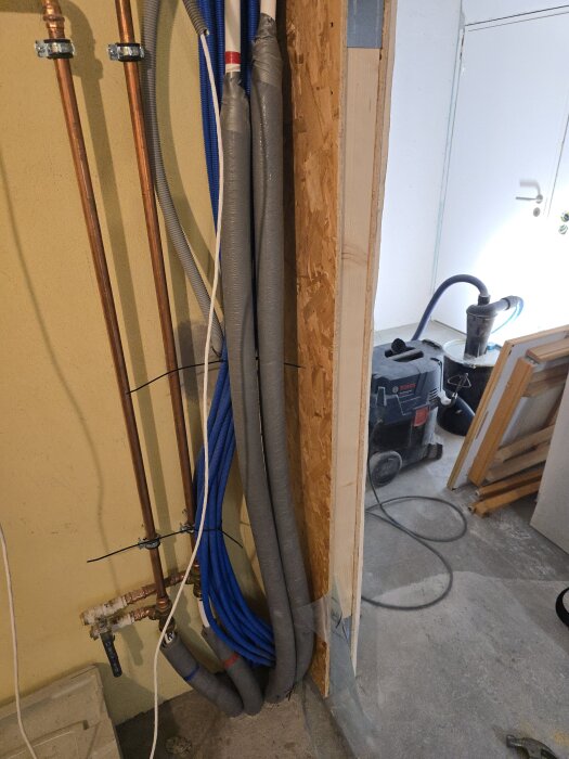 Rör och flexrör monterade längs en vägg i ett hobbyrum/gym, med OSB-skiva och diverse verktyg i bakgrunden.