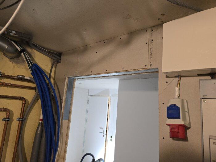 Vägg genom ett rum med synliga rör, kablar och ett eluttag, visar pågående arbete med elinstallationer och väggbeklädnad.