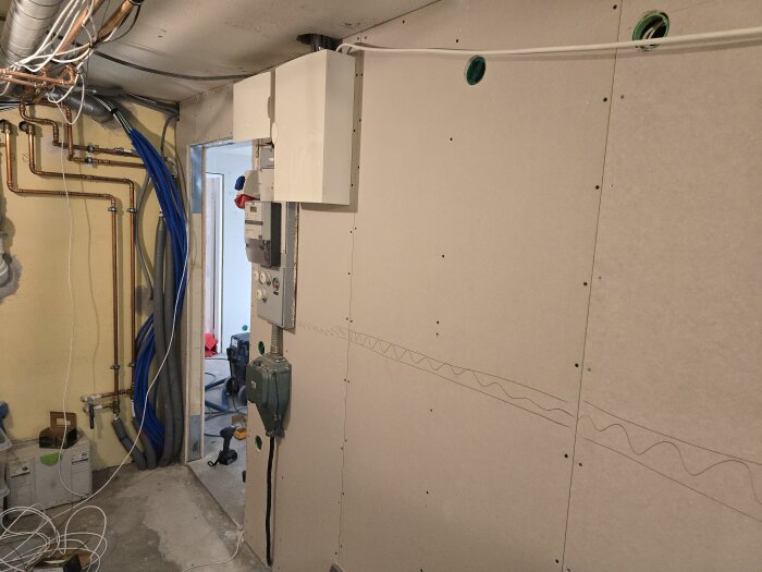 Vägg med osb-skivor och gips i hobbyrum, rörsynliga till vänster. Elinstallationer och flexrör. Renovering pågår med verktyg och kablar på golvet.