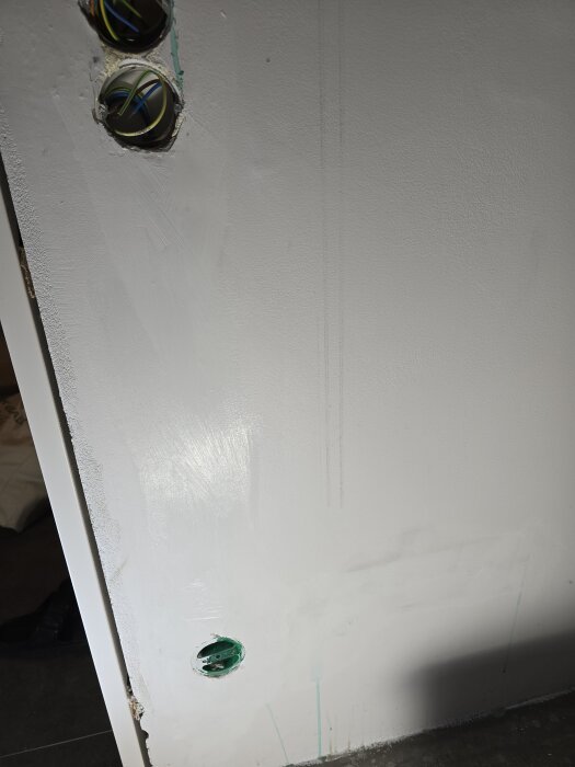 Vägg med gips och eluttag, några synliga kablar i ett övre hål. Nymålad yta med spackel förberedd för fortsatt målning.