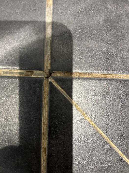 Kakel med sliten fogmassa, nära en avloppsbrunn i badrum. Skuggor syns på plattorna. Fråga om brunnens skick och saknat våtrumsintyg.