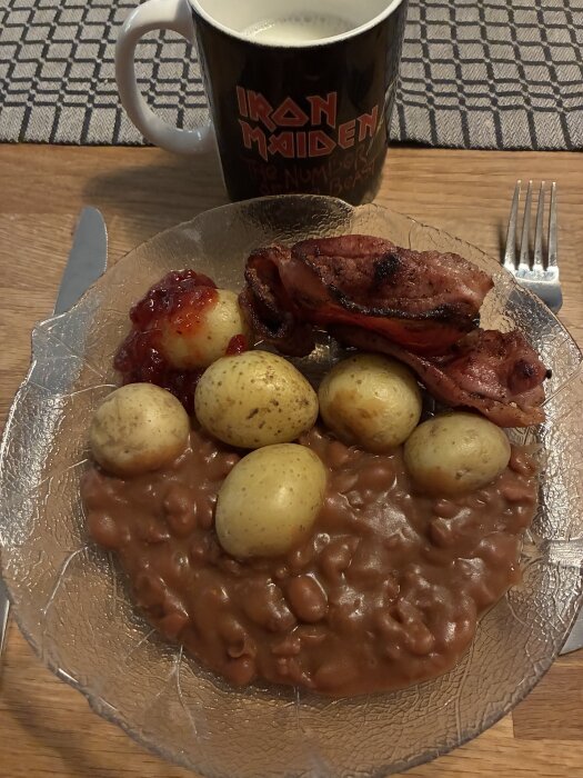 Tallrik med bruna bönor, stekt fläsk, potatis och lingonsylt, samt en Iron Maiden-mugg på ett dukat bord.