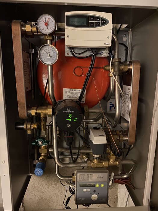 Värmeväxlare i källaren med tryckmätare, rör och elektroniska kontroller; del av ett vattendrivet golvvärmesystem.