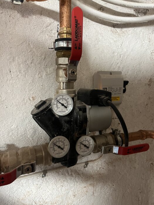 Laddomat-enhet med tre temperaturmätare och kopparrör, ansluten till ett värmesystem i ett hus.