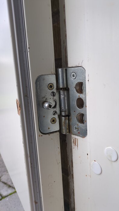 Närbild på ett gångjärn på en ytterdörr som visar skruvhål och slitage på karm och dörrblad.