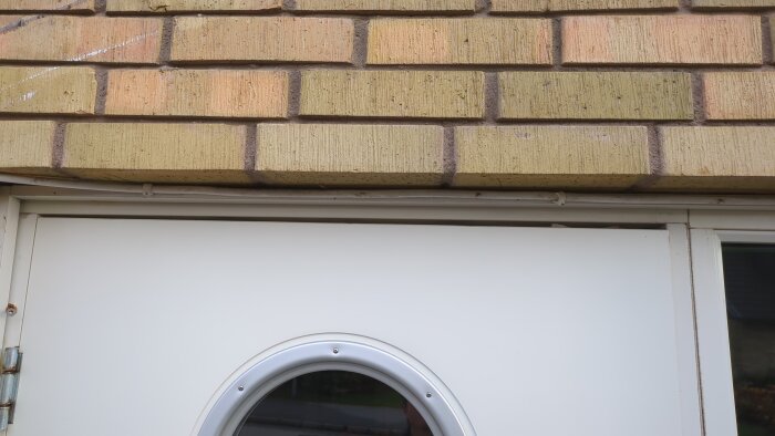 Ytterdörr med rund fönsterruta, tegelvägg och tätningslist vid dörrkarmens överdel, visar justeringsproblem.