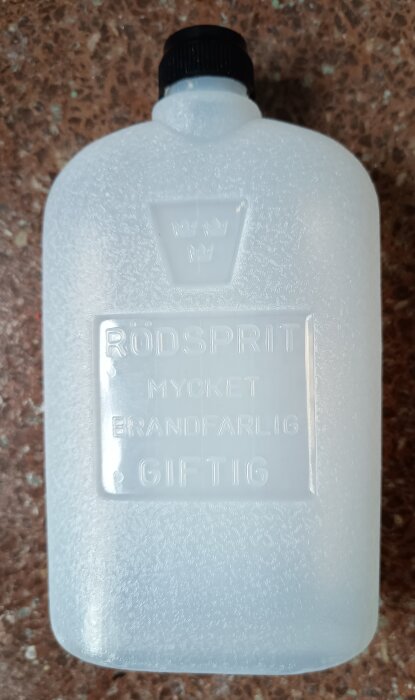 Flaska med etiketttext "Rödsprit", indikerar brandfarlighet och giftighet, placerad på brun yta. Används ofta i enmanskök.