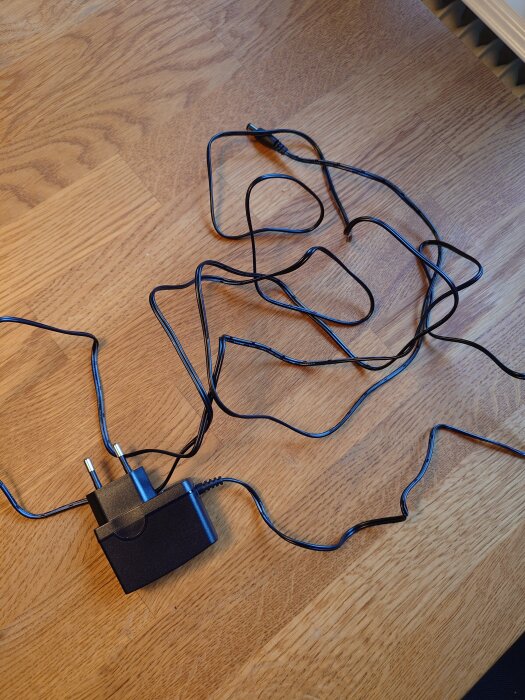 Svart omvandlare med sladd liggande på ett träbord. Sladden ser ut att vara cirka tre meter lång och är ihoptrasslad.