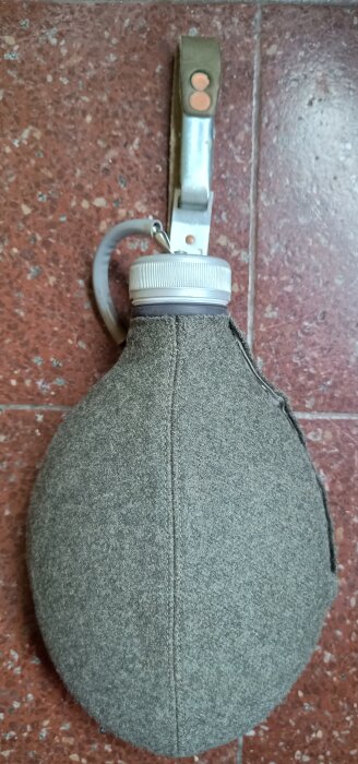 Aluminiumbeklädd vattenflaska med grå filtöverdrag och bärrem, ligger på ett rött kakelgolv.