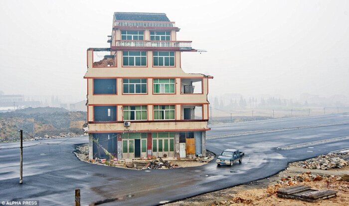 Ett flerfamiljshus står kvar mitt på en nybyggd väg. En bil är i närheten, dimmig bakgrund.