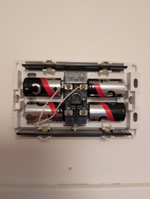 Ett väggmonterat batterifack med fyra C-batterier, kopplade med vita kablar, monterade på en vit vägg.