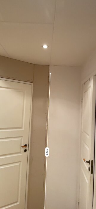 Väggar möts i en vinkel under ett tak med infälld belysning och dörrar på båda sidor, mätt höjd 2,43 m.