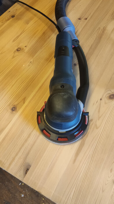 En elektrisk slipmaskin placerad på ett träbord, ansluten till en slang för dammuppsamling.
