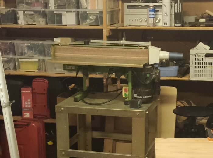 En bandslipmaskin står på ett arbetsbord i en verkstad. Bakom syns hyllor med plastlådor och verktyg.