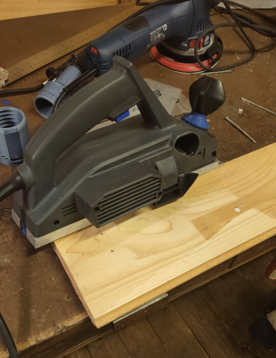 Elektrisk handhyvel på ett träbord, i närheten av en slipmaskin och dammsugaradaptrar. Verktygen är placerade på en arbetsbänk i en verkstad.