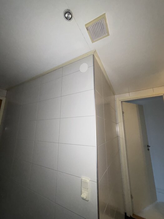 Tak och vägg i badrum med ventilationstrumma och en rund lampa syns i hörnet. Väggkontroll på kaklad vägg.