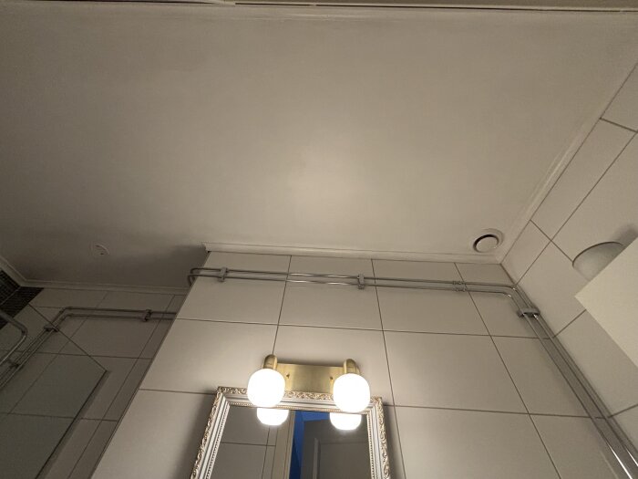 Badrumstak med synliga rör och ventilationsöppning, spegel med belysning under. Vitt kakel på väggarna.