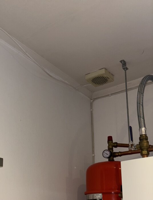 En ventilationsfläkt i dåligt skick är installerad i taket på ett rum med rör och en röd trycktank i förgrunden.