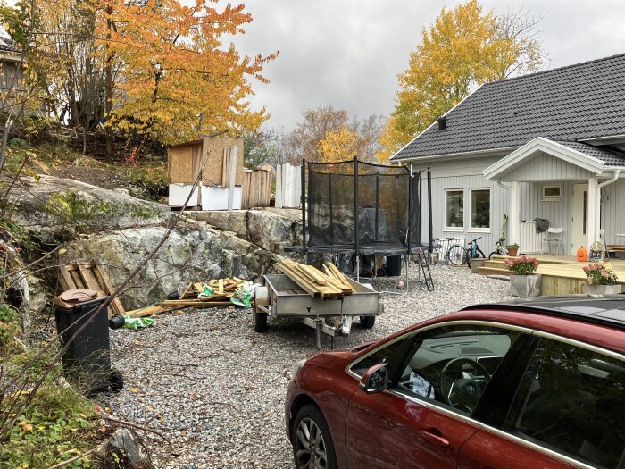 Grusad uppfart framför ett nybyggt hus med berg till vänster, bil, släpvagn och en studsmatta. Höstfärger på träd i bakgrunden.