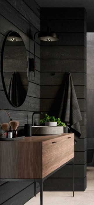 Svartmålade liggande träbrädor på vägg i badrum med rund spegel, svarta handdukar och trähylla med växt.