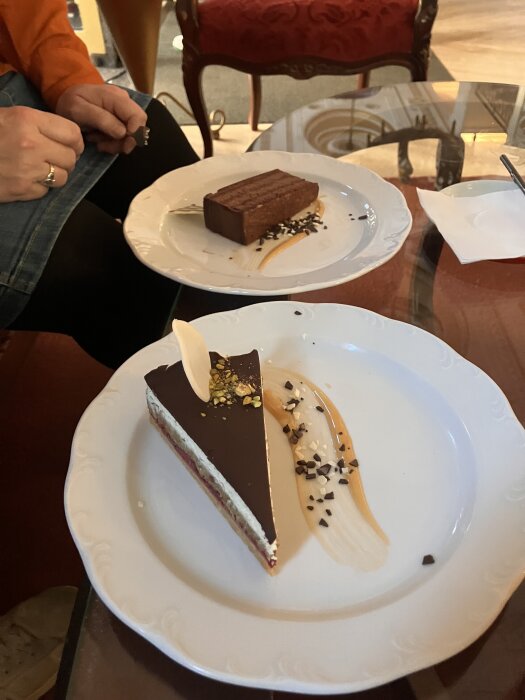 Två desserttårtor på tallrikar, den främsta med chokladtäcke och pistagenötter, den andra en rektangulär tårta. En person håller i en bit choklad.