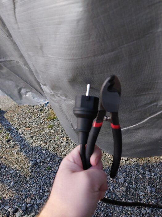 En hand håller en kabel med rak kontakt och en avbitartång framför en presenning utomhus.
