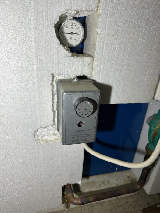 Temperaturmätare och inställningspanel i värmesystemet under fritidshuset, del av pannan och kopparrör synliga.