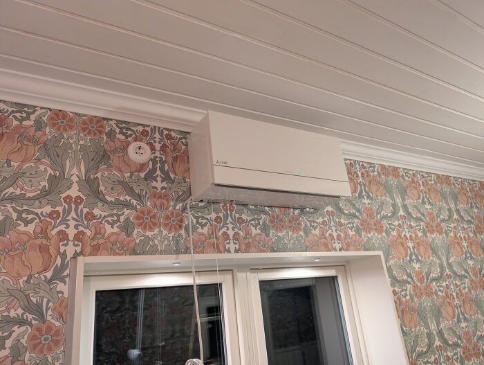 Väggmonterad luftvärmepump ovanför ett fönster mot blommig tapet och vitmålat innertak.