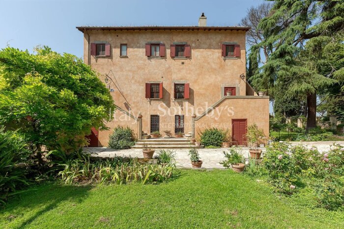 Charmig villa i sandfärgad puts med röda fönsterluckor omgiven av grönskande trädgård och några stora träd i bakgrunden.