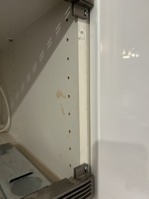 Sidopanel i skåp med flera små hål och skruvbeslag synliga i nedre och övre hörn.