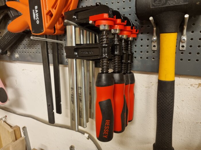 Fyra nya röda och svarta Bessey-tvingar hänger på en verktygstavla bredvid andra verktyg som används i byggen, som en hammare.