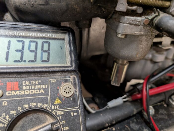 Multimeter visar 13.98V, bredvid en del av en motor där kablar är anslutna.