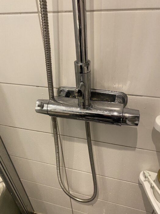 Duschblandare i krom monterad på vit kakelvägg med duschslang hängande nedåt.