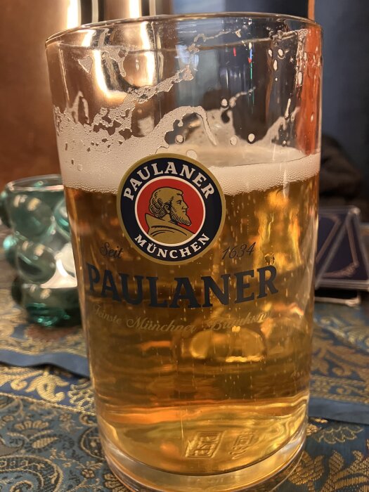 Ett glas med öl från Paulaner på ett bord med blå och guldfärgad duk i bakgrunden.