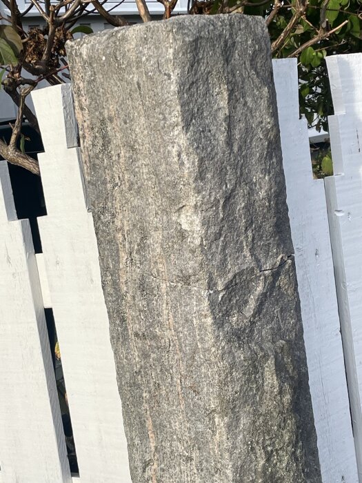 Sprucken stenstolpe vid staket med synlig spricka vid metallinfästning, omgiven av vita träplankor och grönska.
