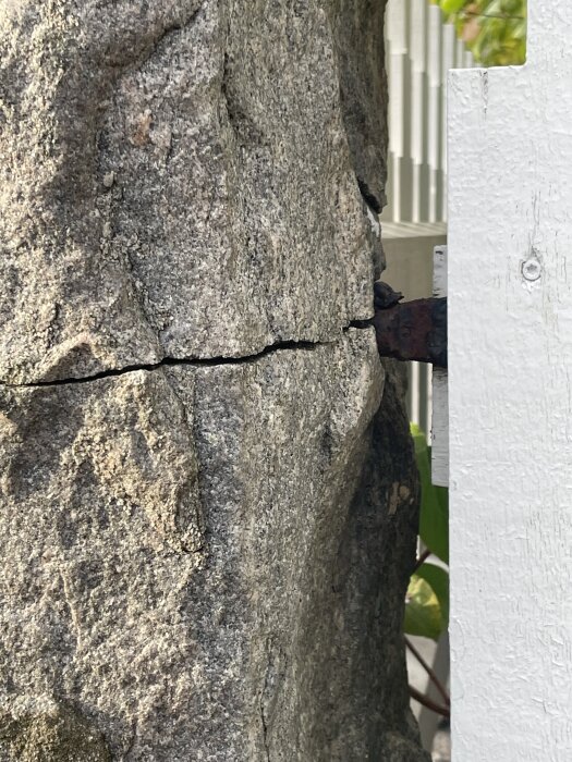 Sprucken stenstolpe vid metallinfästning i ett staket, omgiven av trä och grönska.