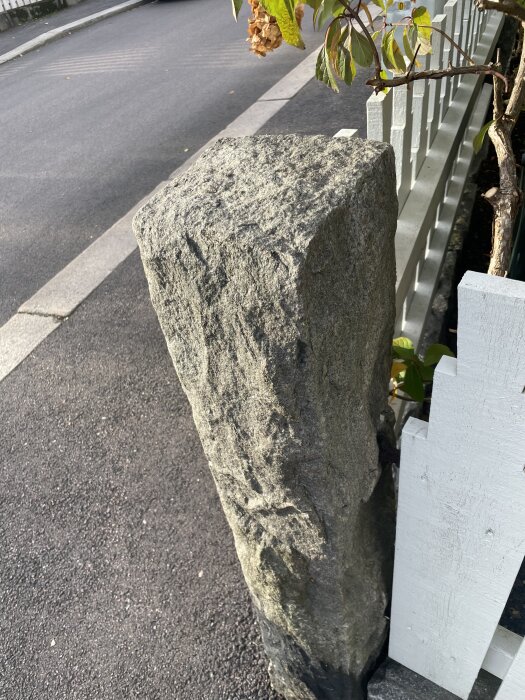 Sprucken stenstolpe vid metallinfästning intill vitt staket, omgiven av asfalt och växtlighet.