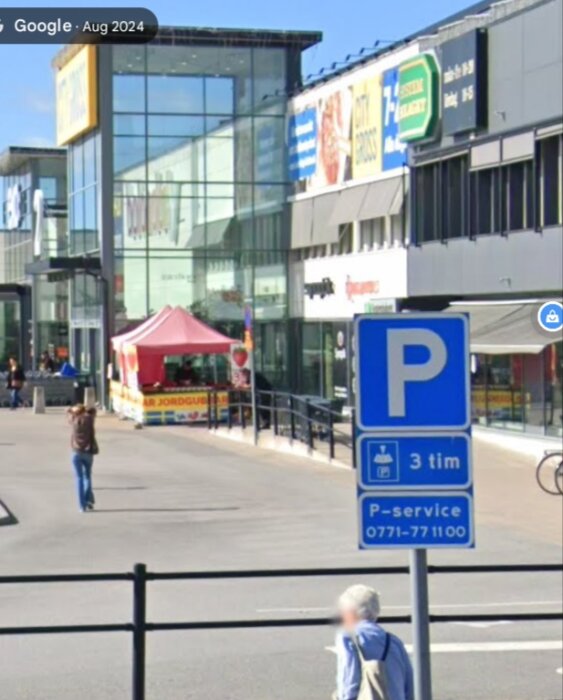 Parkeringsskylt vid stormarknadsområde med text om P-skiva och tidsbegränsning. Bakom syns butiker, en cykel och en man som går.