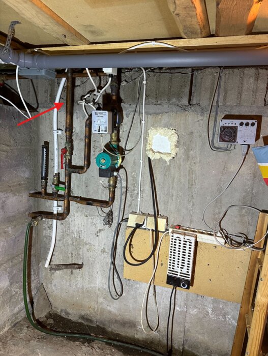 Rörsystem i en krypgrund med vedeldad värmepanna, två pumpar och en pil ritad på ett rör. Väggmonterad utrustning och kablar synliga.