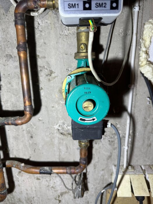 En grön cirkulationspump monterad på rör i en krypgrund, med elektronik märkt "SM1" och "SM2" ovanför.