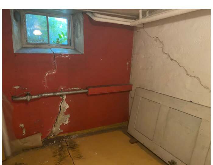 Sprickor i en källarvägg med rött och vitt, mellanvägg till höger, rör under ett litet fönster.