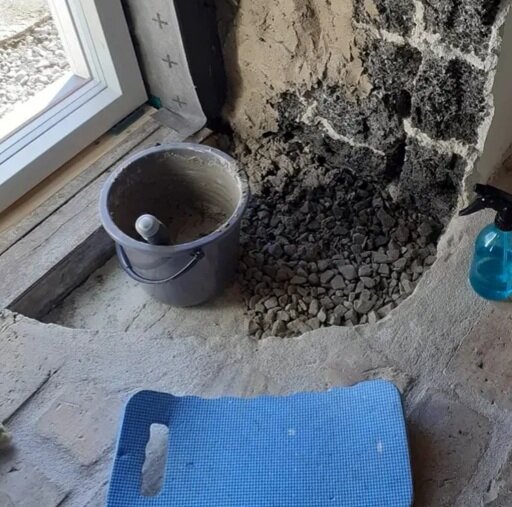 Grus vid en utgrävd vägg i ett ombyggt stall, med en hink och en sprayflaska bredvid. En blå dyna ligger på golvet.