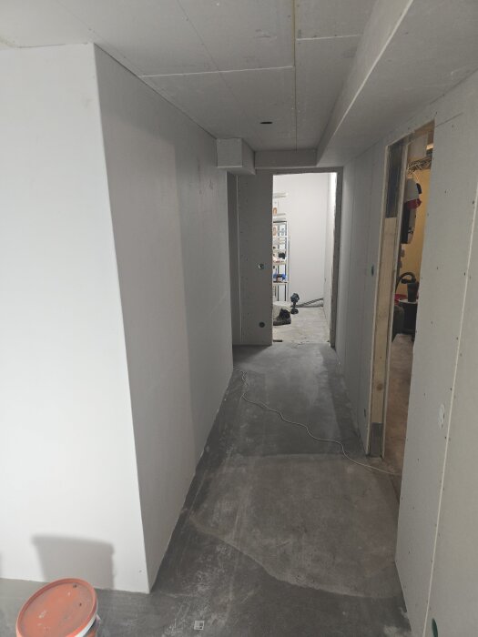 Källargång med nygipsade väggar och tak, påtagligt ren och förberedd för målning. En orange plasthink syns på golvet i förgrunden.