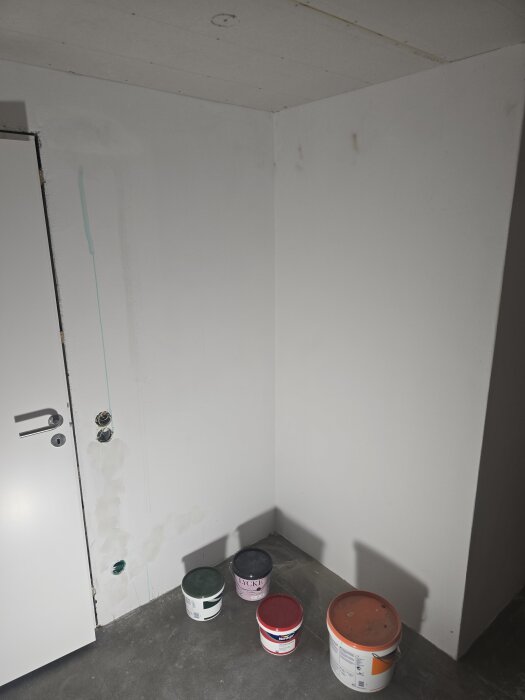 Hörn av ett rum med vitmålade väggar och tak, några målarfärgsburkar på golvet, samt eluttag och dörr till vänster.