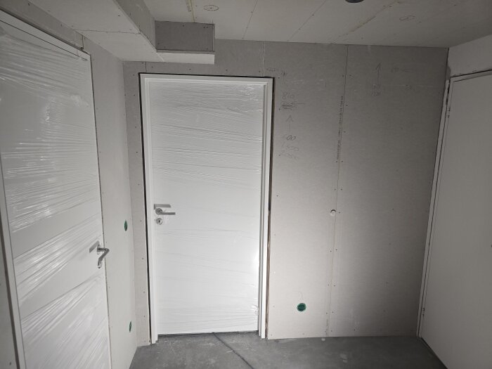 Nya gipsskivor monterade, omålade väggar och dörrar med plastskydd i ett rum färdigt för målarjobb, synliga bygganteckningar på väggen.