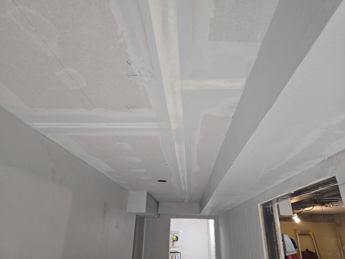 Tak i korridor under renovering med bredspackling och gipsskivor, delvis målat. Öppning i slutet med insyn till rum med arbetslampa och verktyg.